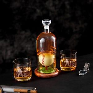Puede incluir: Un juego de decantador de whisky con temática de béisbol. El decantador transparente tiene una pelota de béisbol en su interior, sobre una base de madera con césped artificial. Se incluyen dos vasos a juego con un gráfico de jugador de béisbol y el texto "I'd Hit That".