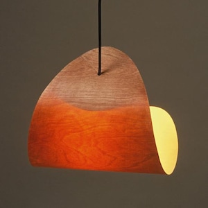 May include: A modern pendant light fixture with a wooden shade. The shade is a curved shape, with a gradient of colors from light brown to orange. A black cord suspends the light from above, and a warm light shines from the side.