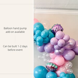 K&ouml;nnte beinhalten: Ein Ballonbogen aus hellblauen, lila und rosafarbenen Ballons. Der Bogen ist mit schillernden Meerjungfrauen-Muschelballons verziert. Der Text "Ballonhandpumpe als Zubeh&ouml;r erh&auml;ltlich" und "Kann 1-2 Tage vor der Veranstaltung aufgebaut werden" ist im Bild sichtbar.