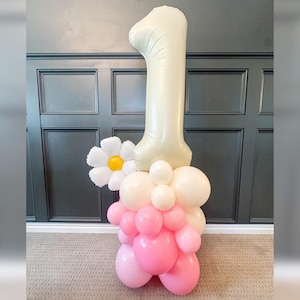 May include: A balloon arrangement for a first birthday celebration. The centerpiece features a large, cream-colored number one balloon, a white daisy, and a cluster of pink and white balloons. The balloons are arranged on a stand.