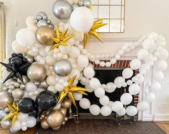 May include: A white fireplace with a balloon garland made of white, gold, silver, and black balloons. The garland is decorated with gold star-shaped balloons.