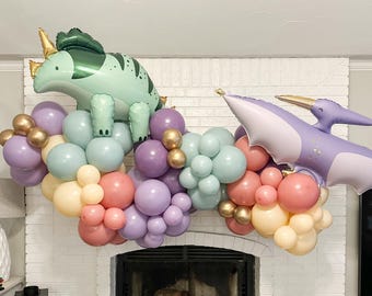 Puede incluir: Un globo de dinosaurio verde y un globo de dinosaurio morado con alas están rodeados de globos de color pastel en tonos de rosa, morado y verde. Los globos están dispuestos en forma de guirnalda.
