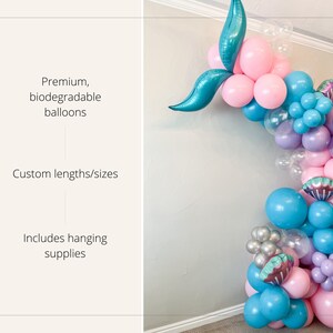 K&ouml;nnte beinhalten: Ein Ballonbogen im Meerjungfrauen-Design aus rosa, blauen und violetten Ballons. Der Bogen ist mit schillernden Muscheln verziert und enth&auml;lt eine t&uuml;rkisfarbene Schwanzflosse. Der Text "Premium, biologisch abbaubare Ballons" ist auf der linken Seite des Bildes sichtbar. Der Text "Individuelle L&auml;ngen/Gr&ouml;&szlig;en" ist unter dem ersten Text sichtbar. Der Text "Enth&auml;lt Aufh&auml;ngebedarf" ist unter dem zweiten Text sichtbar.