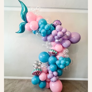 K&ouml;nnte beinhalten: Ein Ballonbogen aus rosa, blauen, violetten und durchsichtigen Ballons. Der Bogen ist mit t&uuml;rkisfarbenen Meerjungfrauen-Schw&auml;nzen und violetten Muscheln verziert.