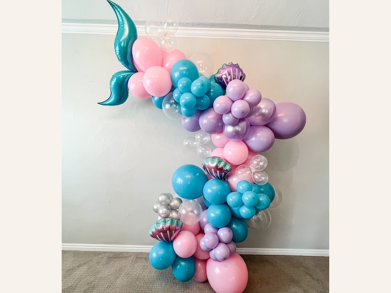 K&ouml;nnte beinhalten: Ein Ballonbogen aus rosa, blauen, violetten und durchsichtigen Ballons. Der Bogen ist mit t&uuml;rkisfarbenen Meerjungfrauen-Schw&auml;nzen und violetten Muscheln verziert.