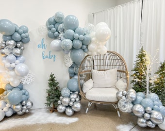 May include: A white wicker egg chair with a white cushion sits in front of a backdrop decorated with blue, white, and silver balloons. The balloons are arranged in an arch shape and include clear balloons with snowflake confetti. The backdrop also features a white sign with the text "oh baby" in blue lettering.