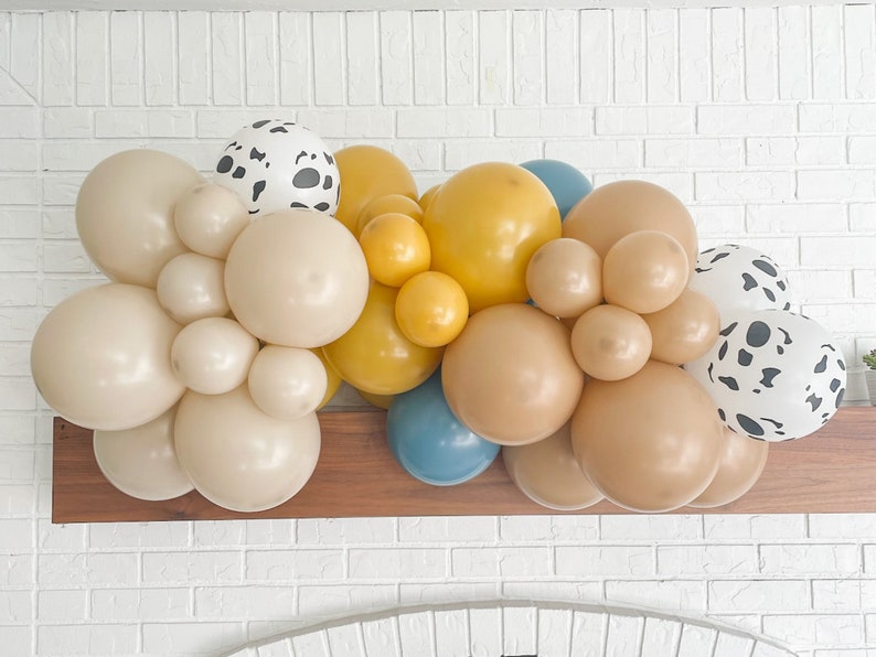 May include: A balloon garland featuring a mix of colors, including beige, yellow, blue, and brown. Some balloons have a dalmatian print. The balloons are arranged in a cluster, suitable for a party or event decoration.