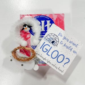 May include: A festive winter-themed collection featuring a penguin ornament with a fluffy white trim, a string of copper-colored fairy lights, and a card that reads "Do you want to build an IGLOO?" with the text "even brought lights and my friend".