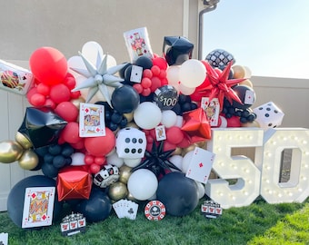 Puede incluir: Un letrero blanco iluminado '50' con bombillas, rodeado de una pared de globos rojos, negros y blancos, cartas de juego y dados. Los globos están dispuestos en cascada, creando un telón de fondo festivo para una celebración.