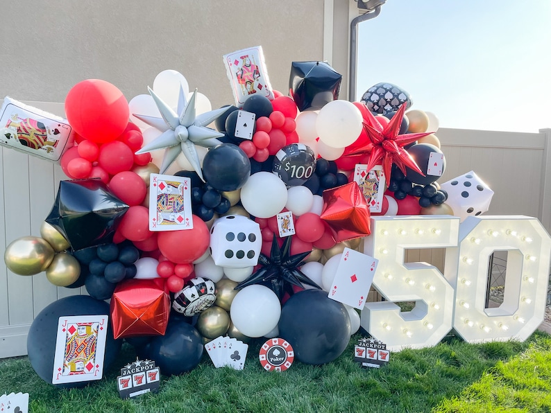 May include: A white, illuminated '50' sign with light bulbs, surrounded by a wall of red, black, and white balloons, playing cards, and dice. The balloons are arranged in a cascading pattern, creating a festive backdrop for a celebration.