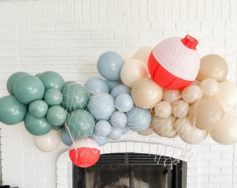 Puede incluir: Un arco de globos hecho de globos azules, verdes y beige con un flotador de pesca rojo y blanco colgando en el centro. El arco está decorado con una red de pesca blanca.
