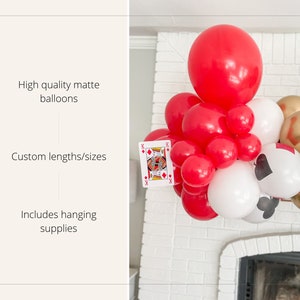 May include: A red, white, and black balloon garland with playing card cutouts. The garland is decorated with red, white, and black balloons, as well as gold and black balloons. The garland is hanging from a white brick wall.
