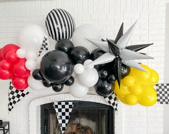 May include: A black, white, and yellow balloon arch with checkered flags. The arch is decorated with balloons in the shape of stars and stripes. The arch is set up in front of a fireplace.