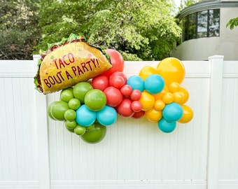 May include: A colorful balloon arch with a large taco-shaped balloon that says "Taco Bout A Party!" The arch is made of green, blue, yellow, and red balloons.