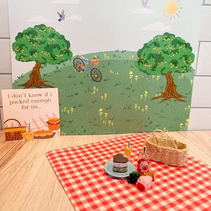 Peut inclure: Une scène de pique-nique miniature avec une nappe à carreaux rouges et blancs, un gâteau en forme de cœur et un panier en osier. Une carte dit : « Je ne sais pas si j'en ai assez emballé pour nous... » Un fond montre un vélo et des arbres.