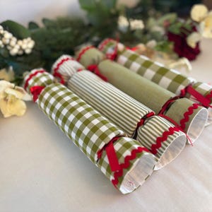 May include: Four Christmas crackers in a row, each with a different design. Two have a green and white check pattern, one has green and white stripes, and one is plain green. They are tied with red ribbon and have a scalloped edge.