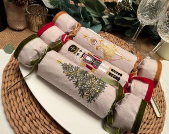 Puede incluir: Un conjunto de petardos navideños decorativos sobre un plato blanco. Cada petardo está envuelto en tela beige y adornado con diseños navideños bordados, incluyendo un árbol de Navidad, un cascanueces y una bailarina. Cintas de terciopelo verde y rojo añaden acentos festivos.