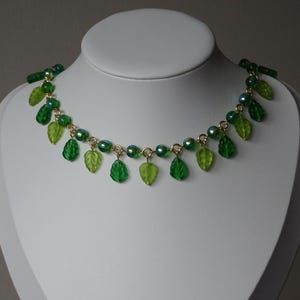 May include: A green necklace with leaf-shaped pendants and iridescent beads. The necklace is displayed on a white mannequin bust. The leaves are various shades of green, and the beads have a shimmering, colorful effect.