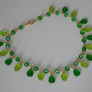 May include: A delicate necklace featuring a gold chain adorned with green and light green leaf-shaped charms and iridescent beads. The necklace has a lobster clasp closure. The leaves and beads are arranged in a cascading pattern.