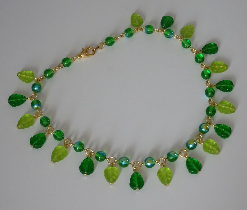 May include: A delicate necklace featuring a gold chain adorned with green and light green leaf-shaped charms and iridescent beads. The necklace has a lobster clasp closure. The leaves and beads are arranged in a cascading pattern.