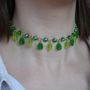 May include: A green beaded necklace with leaf charms. The necklace features a gold chain with green beads and dangling leaf-shaped charms in varying shades of green. The necklace is worn around the neck.