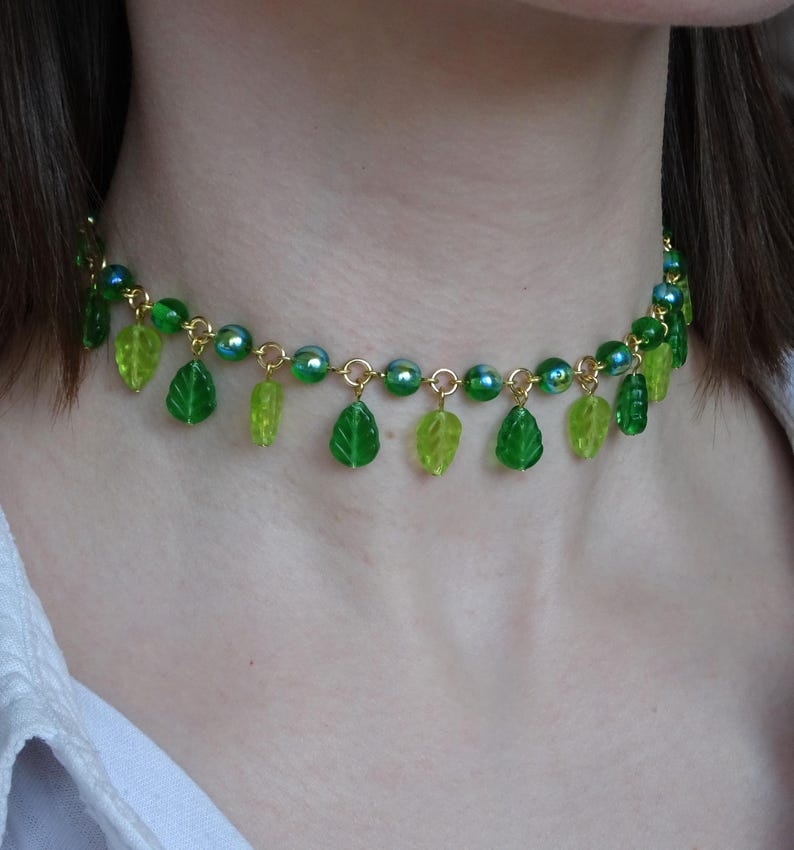 May include: A green beaded necklace with leaf charms. The necklace features a gold chain with green beads and dangling leaf-shaped charms in varying shades of green. The necklace is worn around the neck.