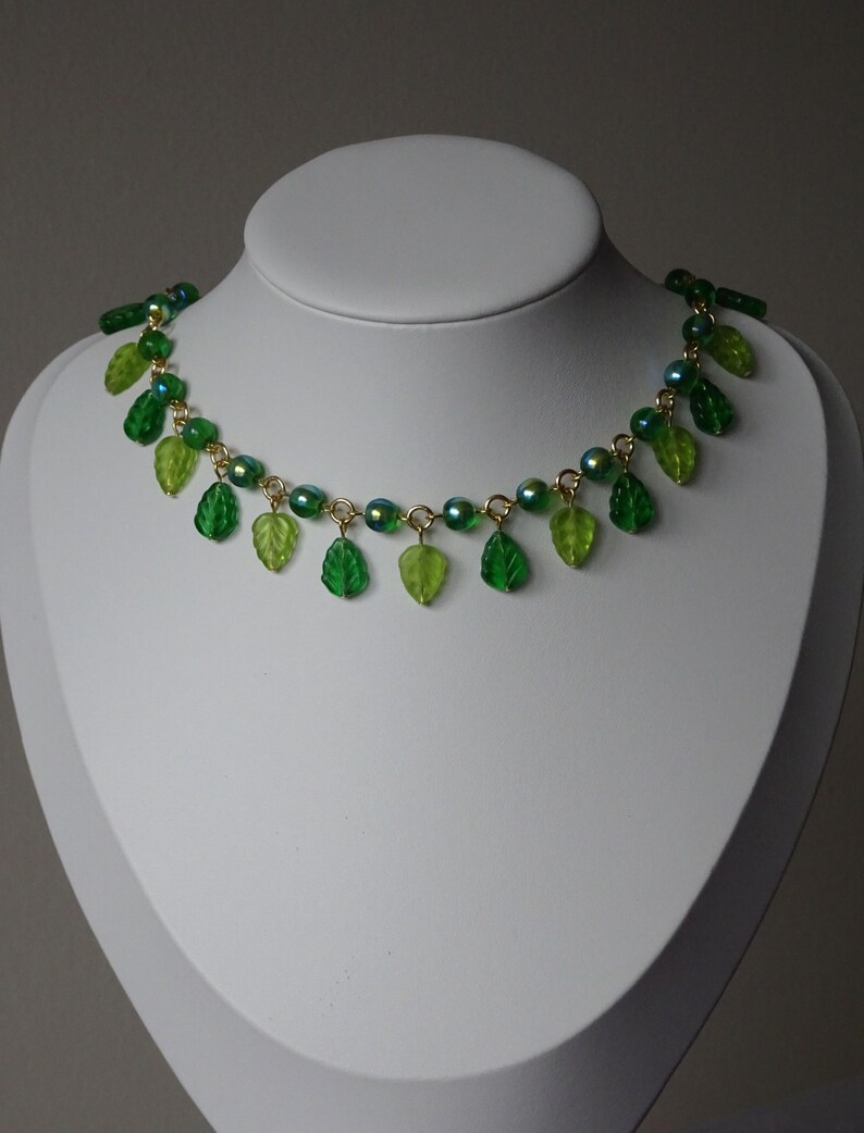 May include: A green necklace with leaf-shaped pendants and iridescent beads. The necklace is displayed on a white mannequin bust. The leaves are various shades of green, and the beads have a shimmering, colorful effect.