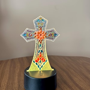 May include: A decorative cross with a colorful, jeweled design. The cross features a central flower design in orange, surrounded by various colored gems. It is mounted on a black base with a light.