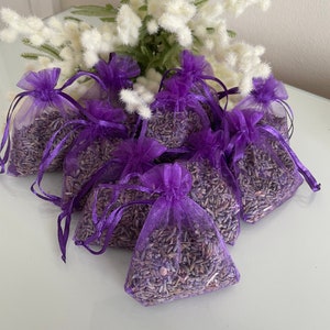 May include: A group of purple organza bags filled with dried lavender. The bags are tied with purple ribbon and are arranged on a white surface.