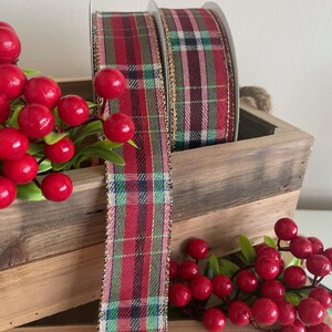 May include: A spool of wired ribbon featuring a red, green, and navy blue plaid pattern with metallic gold edges. The ribbon is displayed alongside clusters of bright red artificial berries with green leaves, resting against a rustic wooden crate.