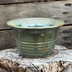 May include: A green ceramic planter with a fluted rim and a brown glaze. The planter is sitting on a weathered tree stump.