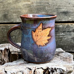 May include: A ceramic mug with a glossy finish featuring a blend of deep blue and purple tones. A textured, light brown maple leaf is embossed on the front surface. The mug has a curved handle and a smooth, rounded rim with a dark brown edge.