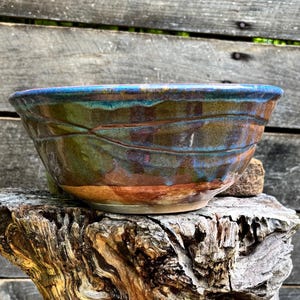 May include: A ceramic bowl with a wide, circular rim and a faceted exterior surface. The glaze features a blend of deep blue, earthy brown, and moss green tones, with subtle horizontal lines etched into the clay body around the entire circumference.