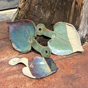 May include: Handmade ceramic leaf-shaped spoon rests in shades of blue, green, and cream. Each rest features a unique leaf vein pattern and a handle with a hole for hanging. The pottery is glazed with a glossy finish.