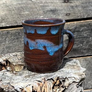 May include: A handmade ceramic mug with a brown base and a blue glaze. The mug has a handle and a textured surface. The blue glaze drips down the sides of the mug, creating a unique design. The mug is sitting on a piece of wood.