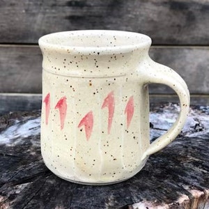 May include: A white ceramic mug with a speckled glaze and pink abstract designs. The mug has a rounded body and a handle.