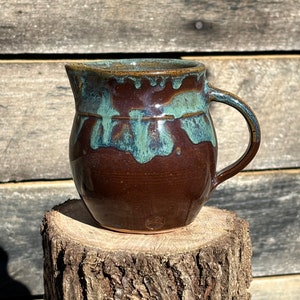 May include: A brown ceramic pitcher with a light blue glaze. The pitcher has a rounded body and a handle. It is sitting on a tree stump.