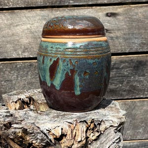 May include: A handcrafted ceramic jar with a lid. The jar features a rich color palette of brown, teal, and bronze. The glaze creates a textured, glossy finish. The jar is cylindrical with a rounded lid, perfect for storing small items.