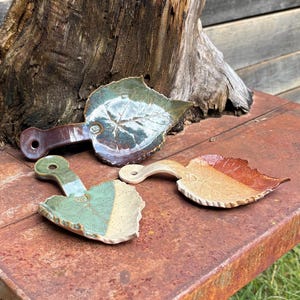 May include: Three ceramic leaf-shaped serving dishes in shades of green, brown, and tan. Each dish has a handle and a detailed leaf vein pattern. The dishes are displayed on a rusty metal surface.
