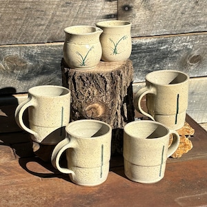 May include: Set of five ceramic mugs with a speckled beige glaze and green accents. The mugs have a tall cylindrical shape and a handle on one side. Two smaller ceramic pitchers with a similar glaze and green accents are also included.
