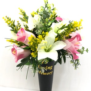 May include: A black plastic flower vase with a gold inscription that reads "In Loving Memory". The vase is filled with a bouquet of pink roses and white lilies with green foliage.