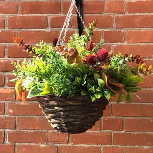 May include: A brown wicker hanging basket filled with artificial greenery, including green leaves, red flowers, and brown branches.