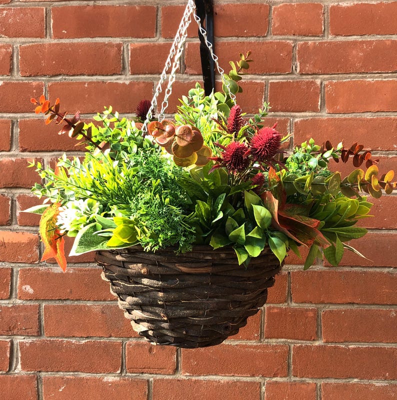 May include: A brown wicker hanging basket filled with artificial greenery, including green leaves, red flowers, and brown branches.