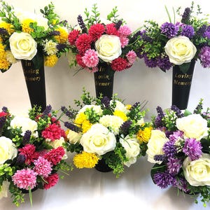 May include: Artificial flower arrangements in black cone vases. The flowers include white roses, yellow pom-poms, and purple lavender. The vases are inscribed with "In Loving Memory" in gold lettering.