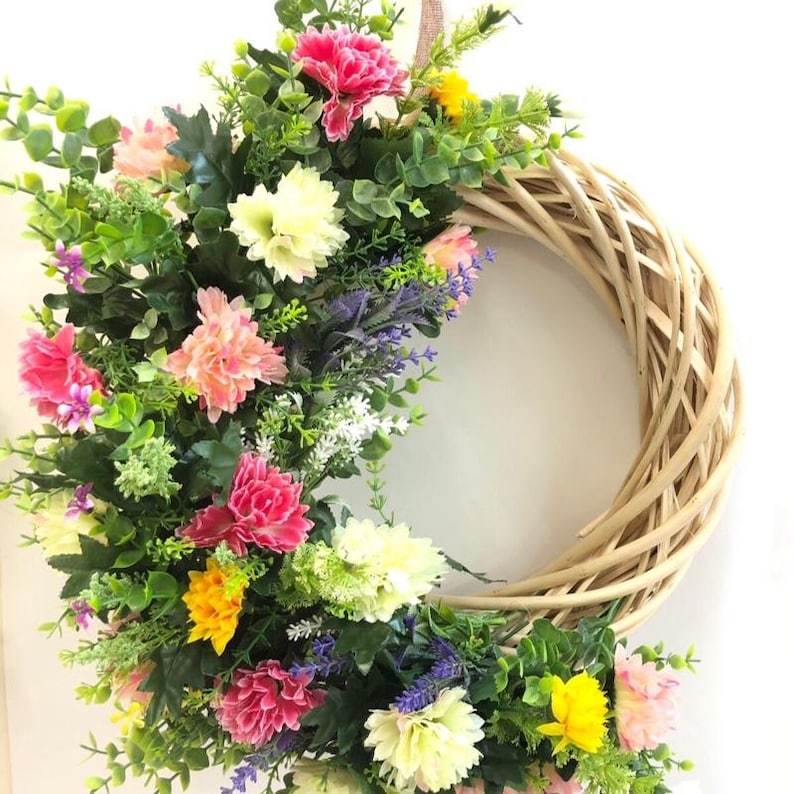 May include: A circular wreath constructed from light brown woven twigs, adorned on one side with an arrangement of artificial flowers in shades of pink, yellow, white, and purple, interspersed with green foliage and small sprigs of lavender.