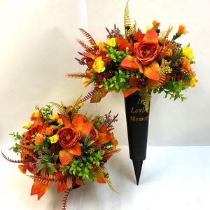 May include: Two floral arrangements featuring orange and yellow roses, green foliage, and small dark berries. One arrangement is set in a black conical grave marker with the gold text In Loving Memory printed on the front, while the other is round.