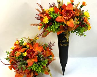 May include: Two floral arrangements featuring orange and yellow roses, green foliage, and small dark berries. One arrangement is set in a black conical grave marker with the gold text In Loving Memory printed on the front, while the other is round.
