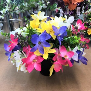 May include: A colourful floral display in a black pot. The arrangement includes artificial flowers in pink, yellow, white, and blue. The flowers are densely packed, creating a full and attractive display. Other floral arrangements are visible in the background.