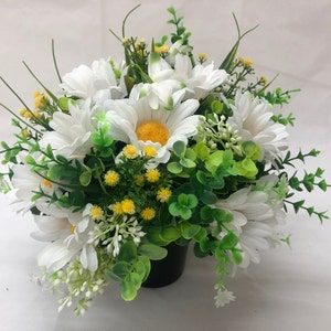 May include: A floral arrangement of white daisies with yellow centres, small yellow flowers, and green foliage. The flowers are arranged in a black pot, creating a vibrant and natural aesthetic.
