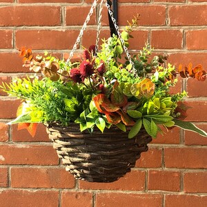 May include: A hanging basket filled with faux greenery and red and orange leaves. The basket is made of woven brown twigs.